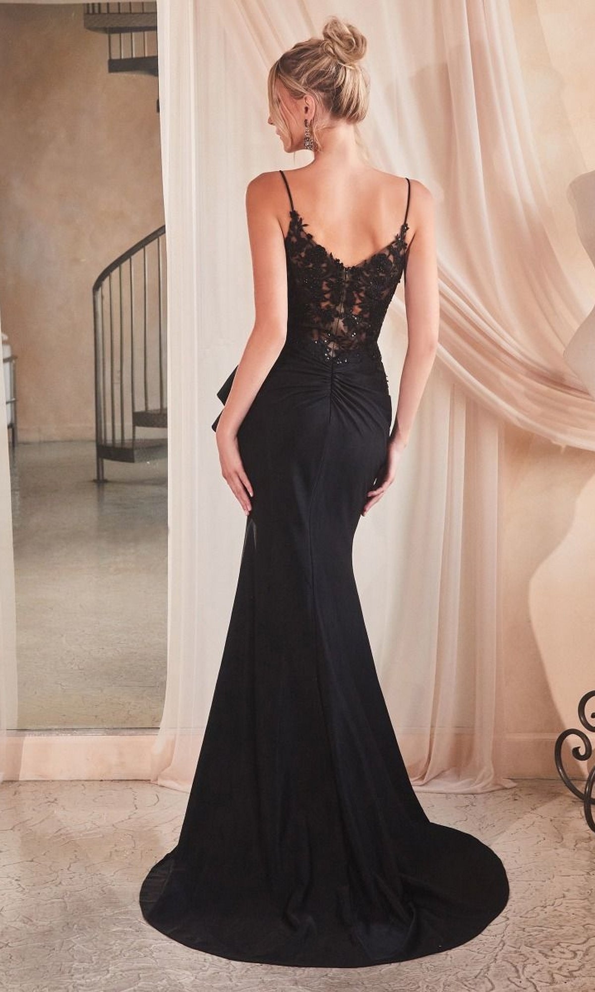 A woman stands indoors near draped curtains and a spiral staircase, facing away from the camera, wearing the Ladivine Long Black Formal Dress KV1100 featuring a lace back detail.